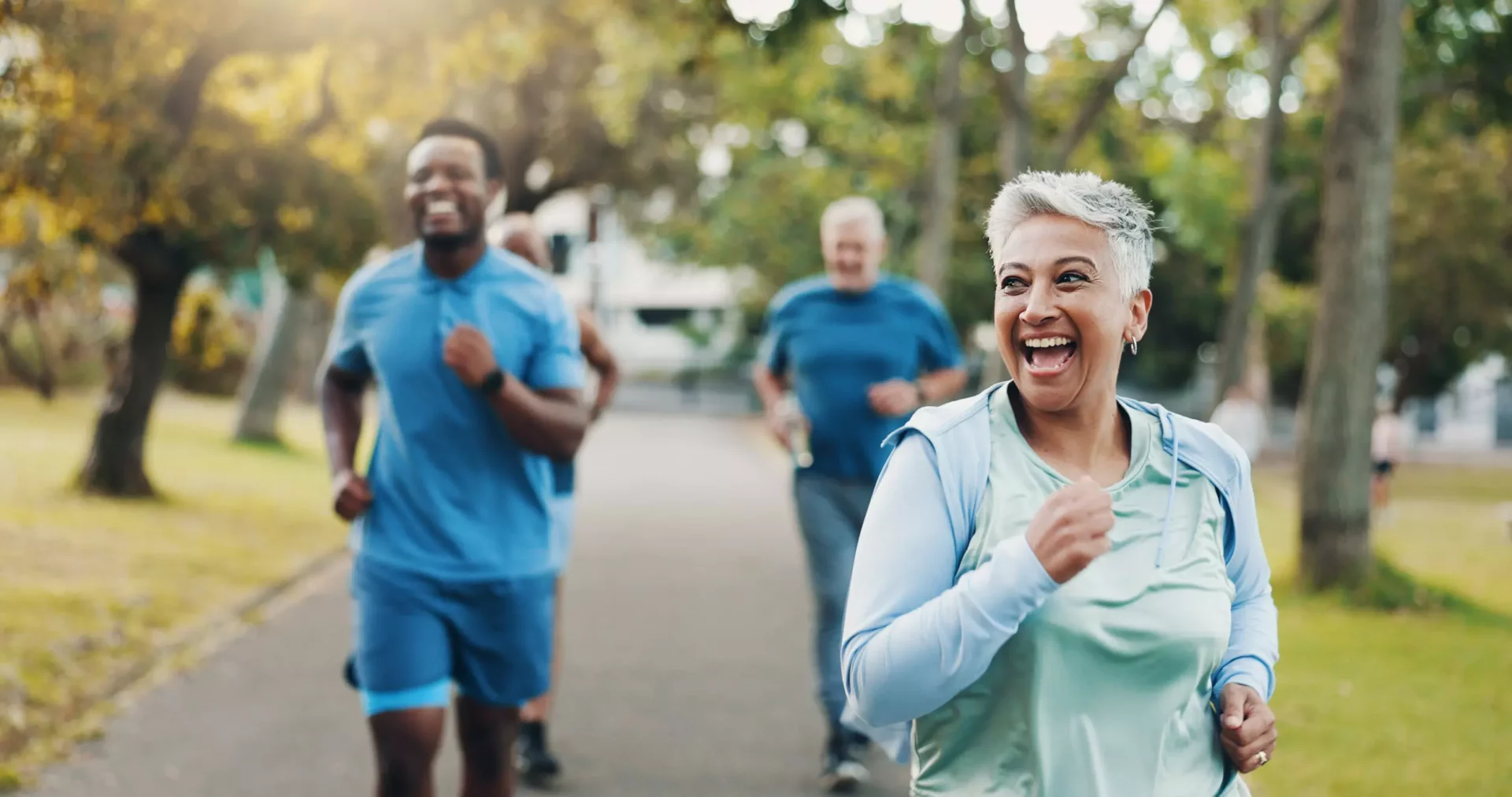 A smiling older woman jogging in a Colorado park, with three people running behind her on a sunny day. The group looks happy and active, surrounded by green trees—enjoying outdoor life and the benefits of good health and Medicare coverage.